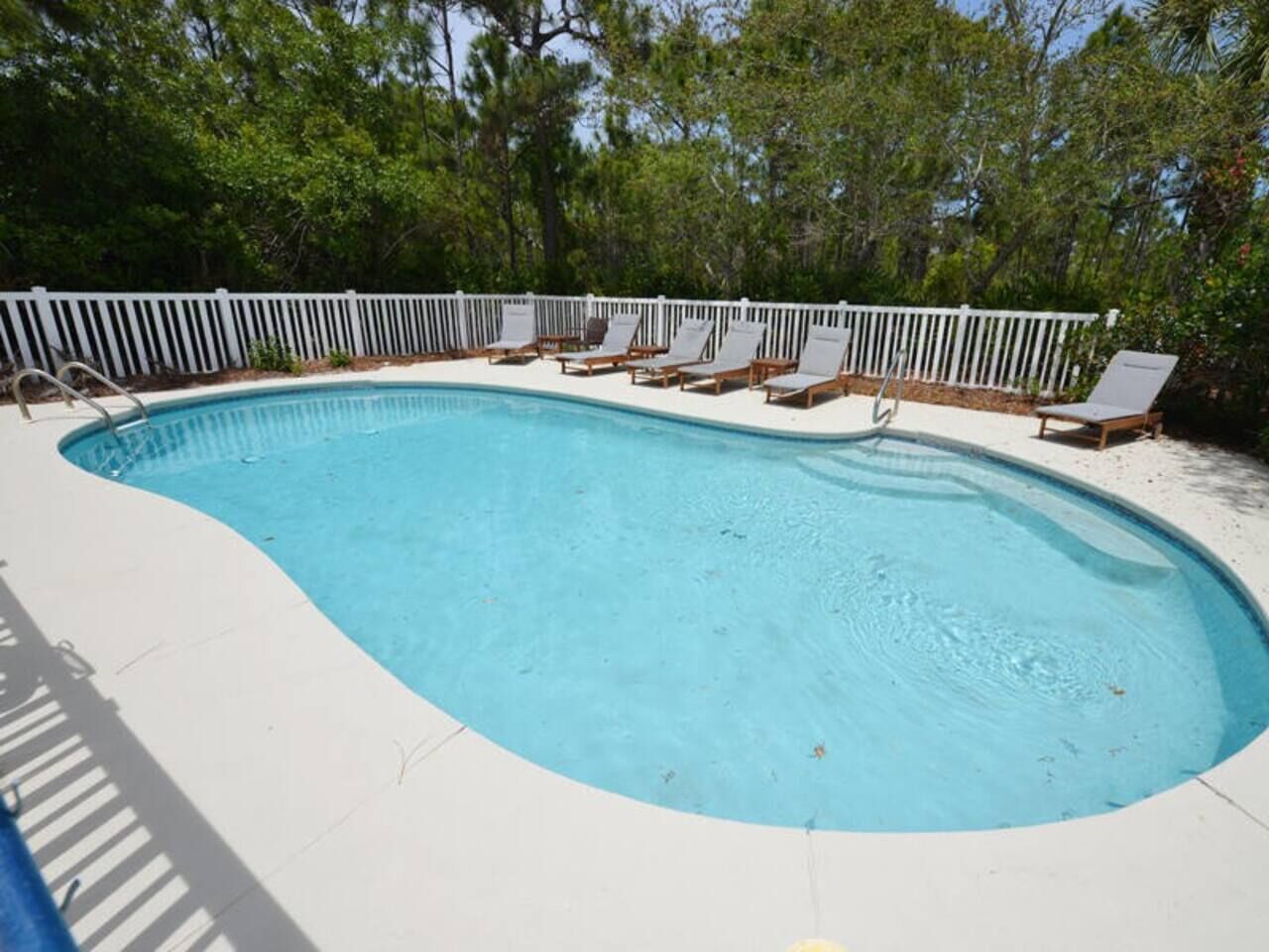 A swimming pool with a white deck, surrounded by a white fence and trees.