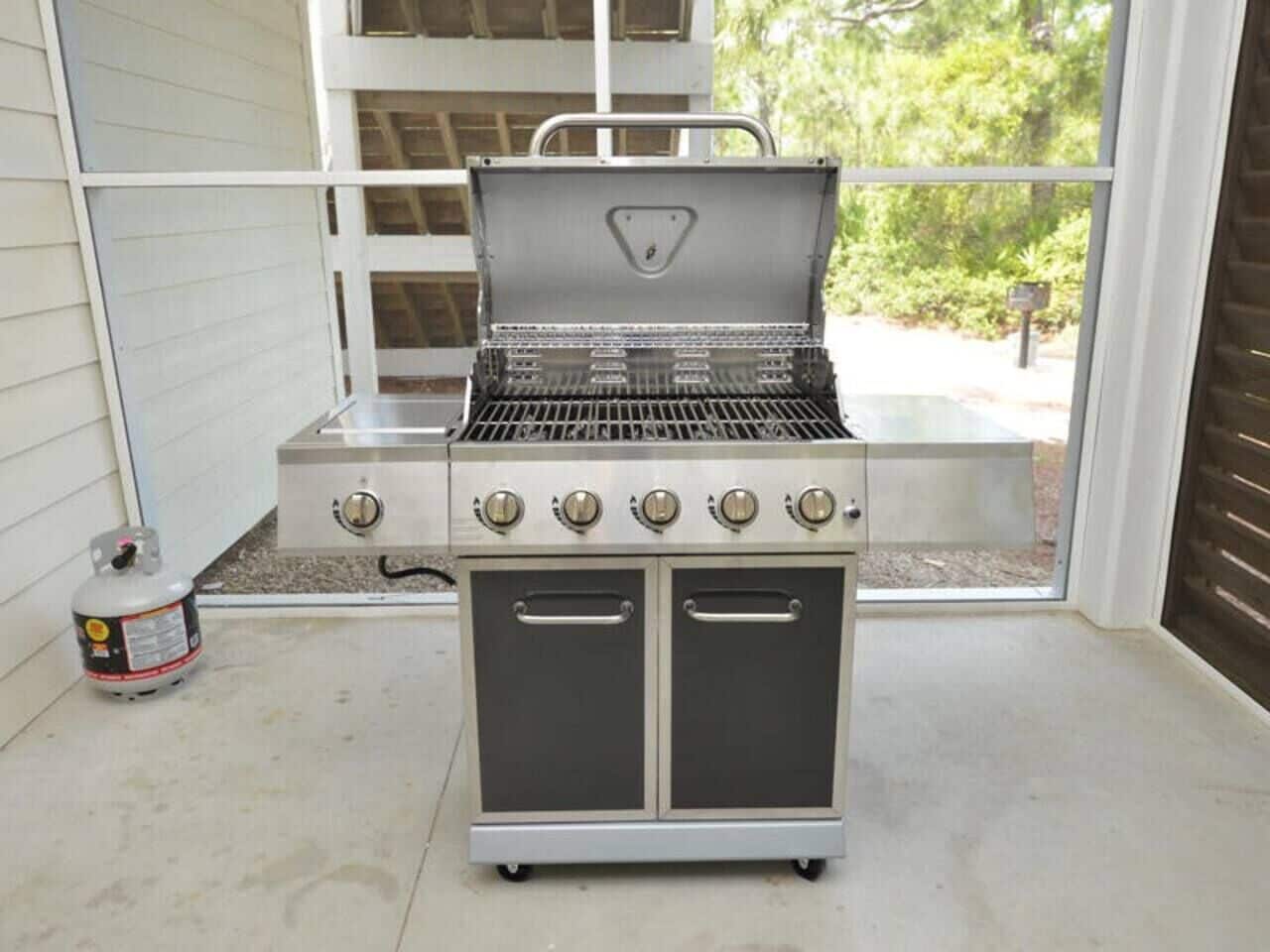 Un barbecue extérieur en acier inoxydable, doté de quatre brûleurs et de deux placards latéraux, installé sur une terrasse en béton avec une fenêtre en arrière-plan.