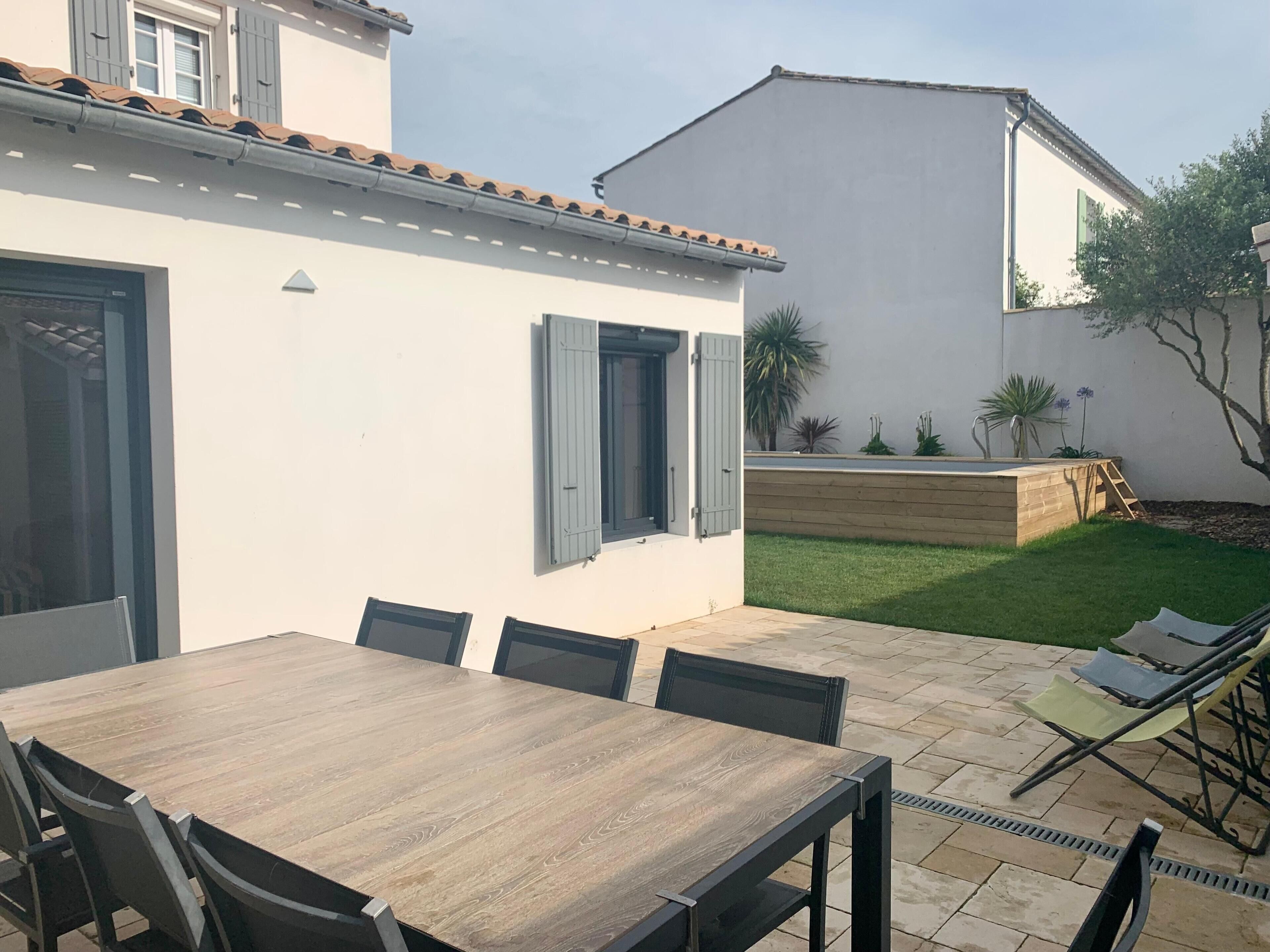 A patio area with a wooden table and chairs, a small garden with plants, and a white building with blue shutters.