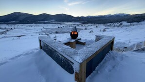 Área externa coberta de neve, com lareira, cercada por uma estrutura de madeira e vista para montanhas ao longe.