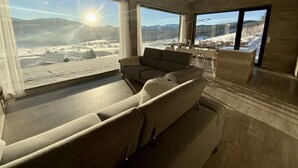 Uma sala de estar ampla com um sofá grande e uma mesa de jantar, com janelas grandes que oferecem vista para uma paisagem nevada.