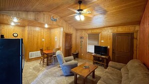 Interior acollidor d'una cabana amb taula de menjador, cadires, sofà, televisió i ventilador de sostre.