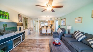 A living room with a ceiling fan, a dining area with a table and chairs, and a kitchen in the background.