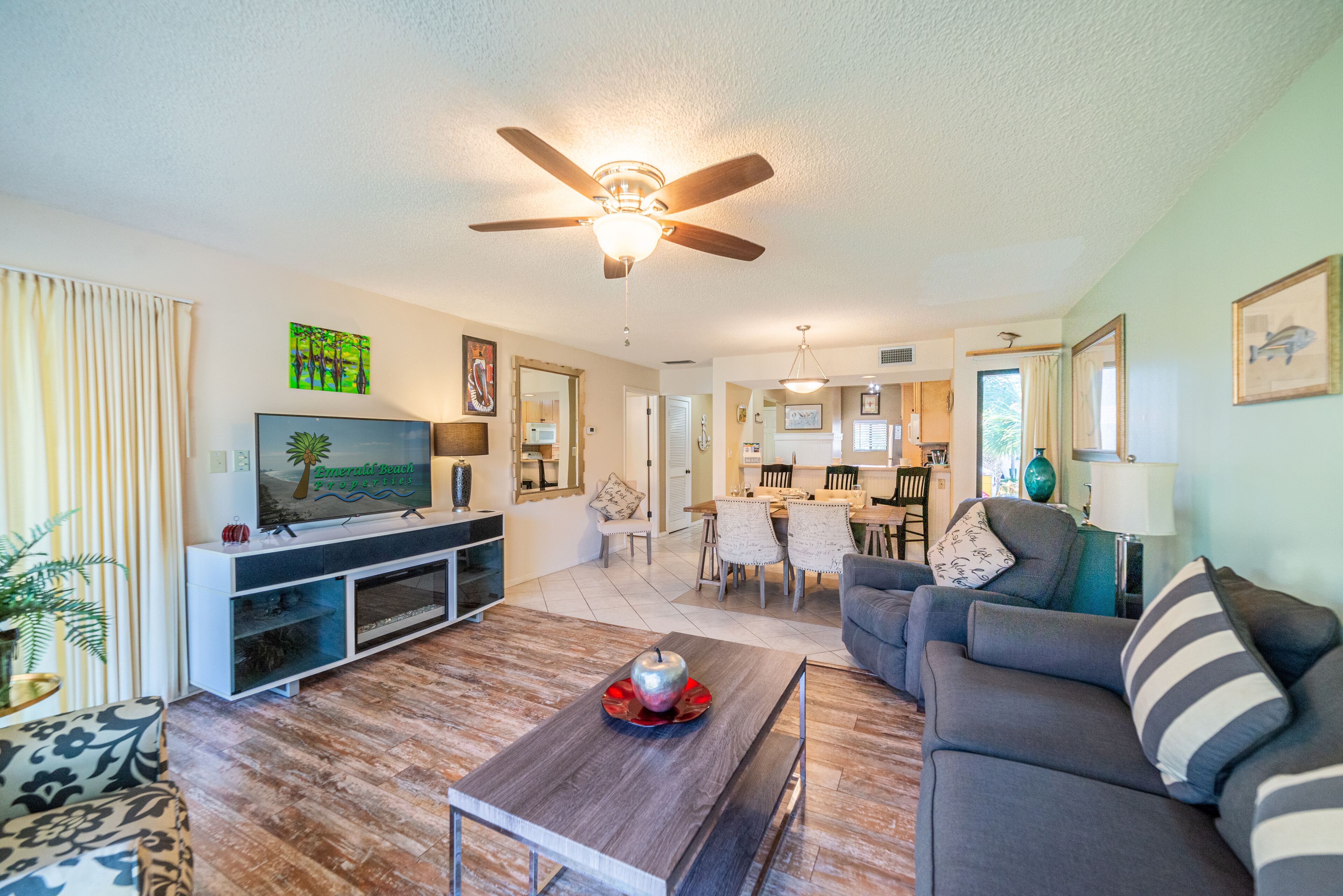 A modern living room with a flat-screen TV, a ceiling fan, and a dining area in the background.