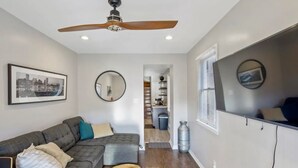 A modern living room with a gray sectional sofa, a ceiling fan, a framed cityscape picture, a round mirror, and a kitchen area in the background. - Co's Corner (Baltimore)