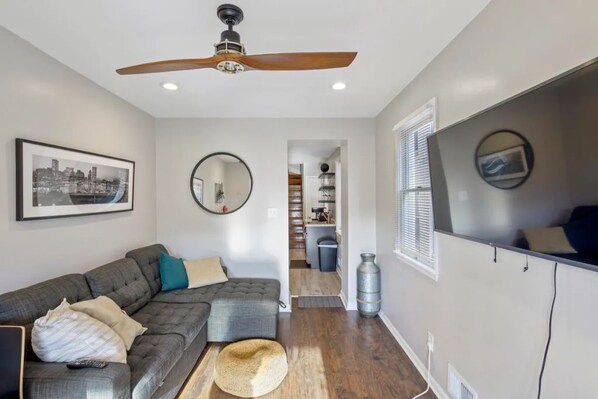 A modern living room with a gray sectional sofa, a ceiling fan, a framed cityscape picture, a round mirror, and a kitchen area in the background. - Co's Corner (Baltimore)