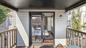 A balcony with a wooden floor, a glass door leading to an outdoor seating area, and a view of a neighboring building. - Co's Corner (Baltimore)