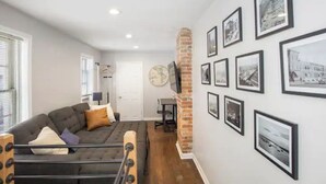 A modern living room with a sofa, a TV, and a brick column. There are multiple framed pictures on the wall. - Co's Corner (Baltimore)