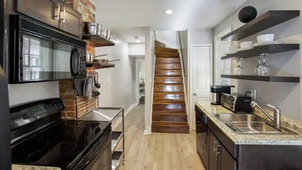 A modern kitchen with a built-in oven, microwave, and stainless steel sink. There is a wooden staircase leading to another room. - Co's Corner (Baltimore)