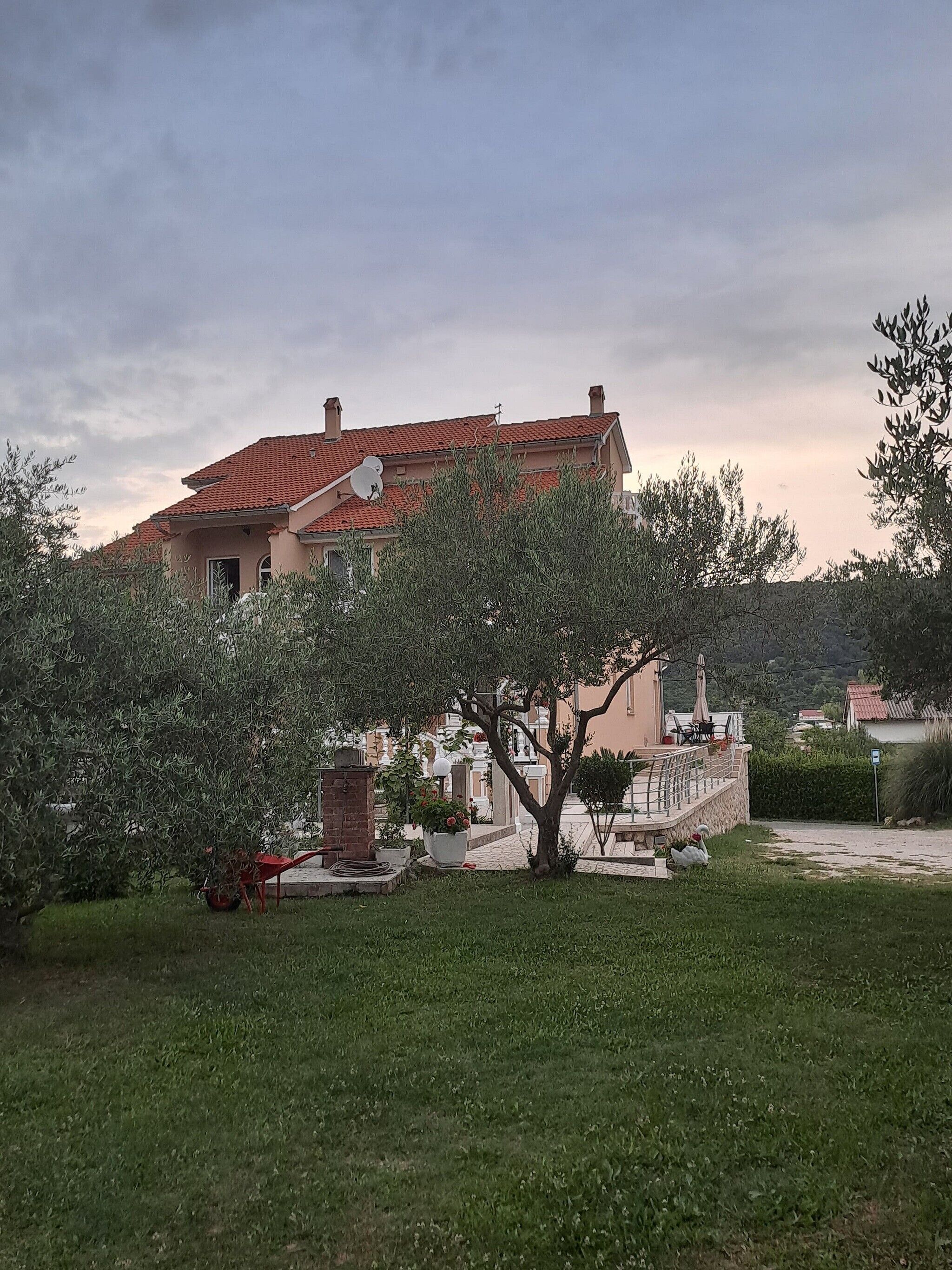 A house with a red-tiled roof, a garden with a tree, and a paved area with outdoor furniture.