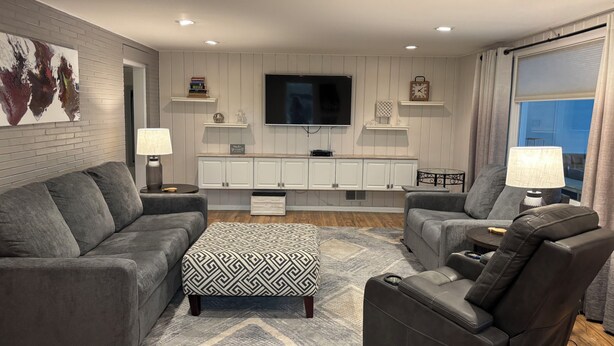Un salon moderne avec deux canapés gris, un pouf à motifs, un fauteuil inclinable en cuir, un téléviseur à écran plat et un meuble de rangement blanc pour les appareils multimédias.
