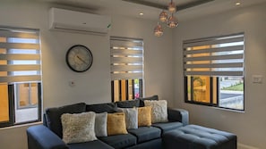 A modern living room with a dark-colored sectional sofa, a round wall clock, and large windows with horizontal blinds.