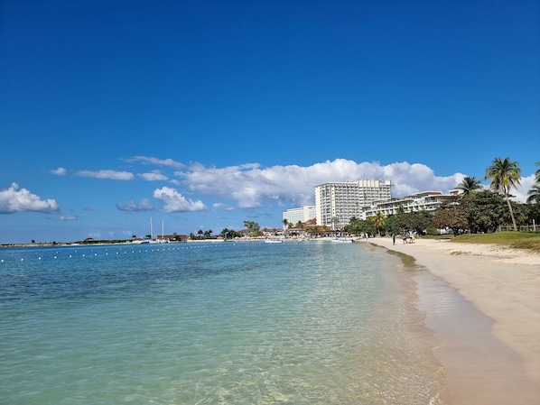 A beach with clear blue water, a sandy shore, and a large hotel building in the background.