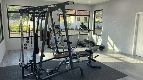 Un gimnasio con diversos equipos de ejercicio, ventanas amplias y vistas al exterior.