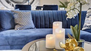 Ein blaues Sofa mit gemusterten Kissen, drei brennende Kerzen und eine Zimmerpflanze mit goldfarbenen Blättern auf einem Glastisch.