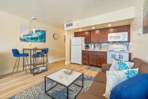 A compact living space with a kitchenette, a dining area with blue chairs, a sofa with patterned cushions, and a marble coffee table.