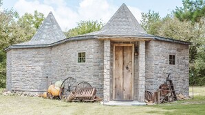 Un edificio in pietra con una porta di legno e due finestre, immerso in un'area erbosa con attrezzature agricole.