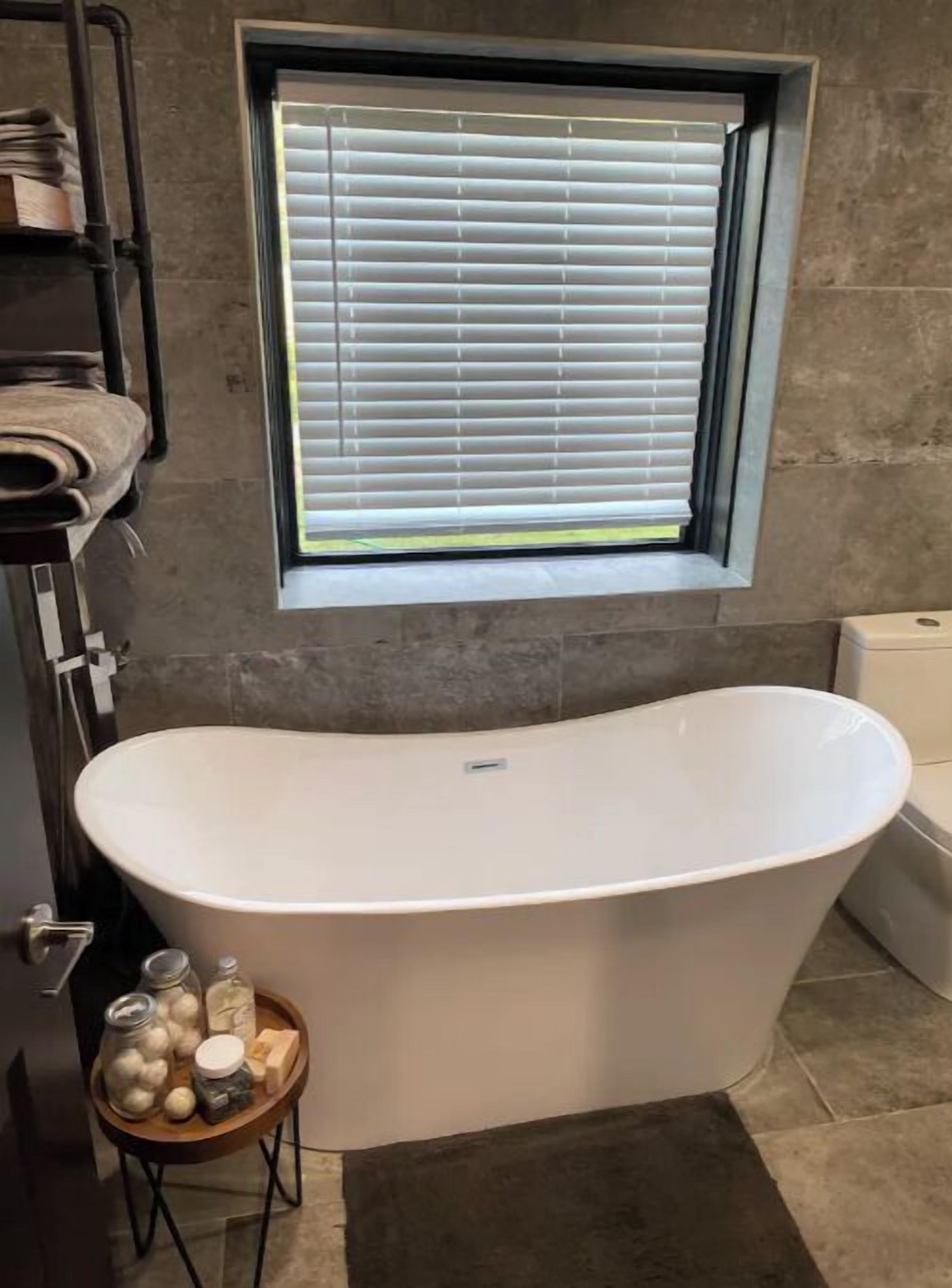 A bathroom with a freestanding bathtub, a window with blinds, a towel rack, and a small table with toiletries.