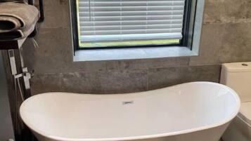 A bathroom with a freestanding bathtub, a window with blinds, a towel rack, and a small table with toiletries.