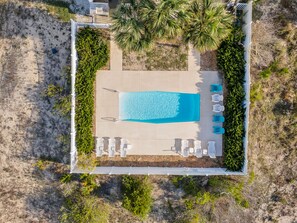 An aerial view of a rectangular swimming pool with a tiled deck, surrounded by white lounge chairs and a few palm trees.