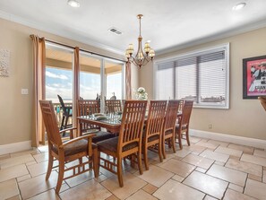 Un coin repas avec une table dressée pour six personnes, des chaises en bois, un lustre et de grandes fenêtres équipées de stores.