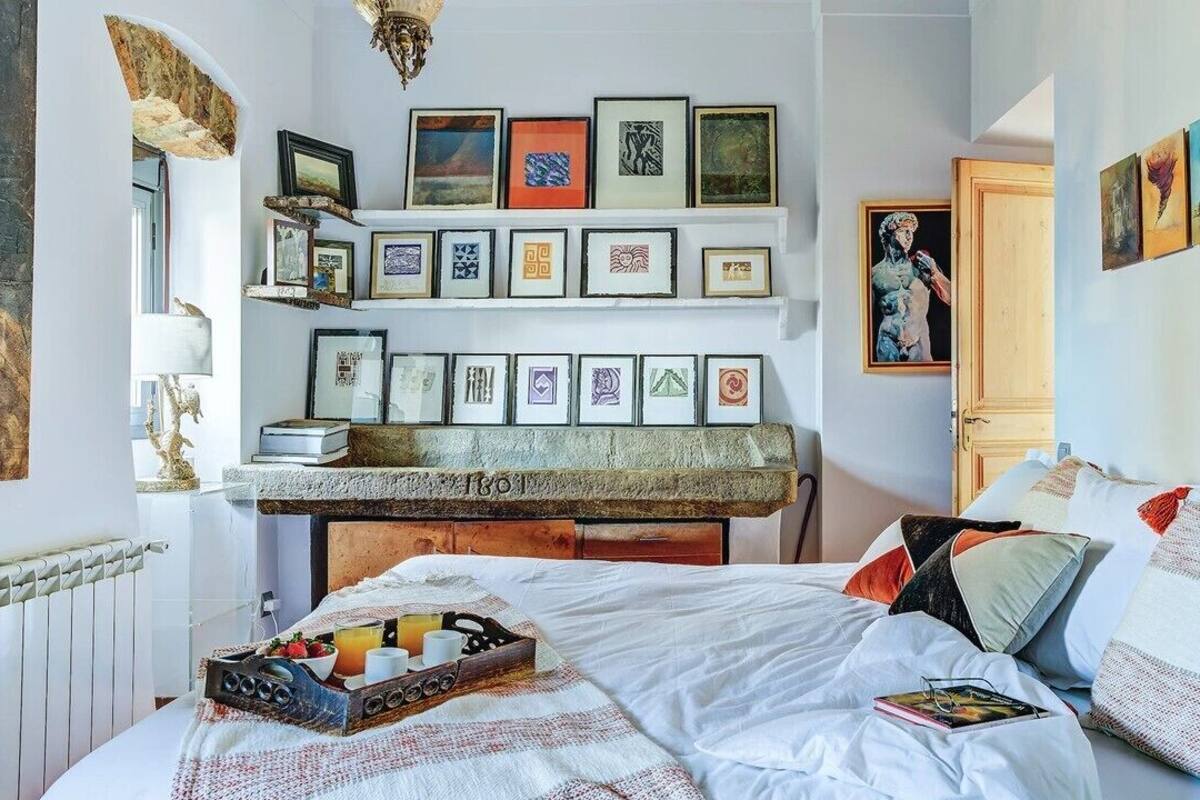 Un dormitorio con cama, mesita de noche con bandeja de desayuno, chimenea y pared decorada con cuadros.