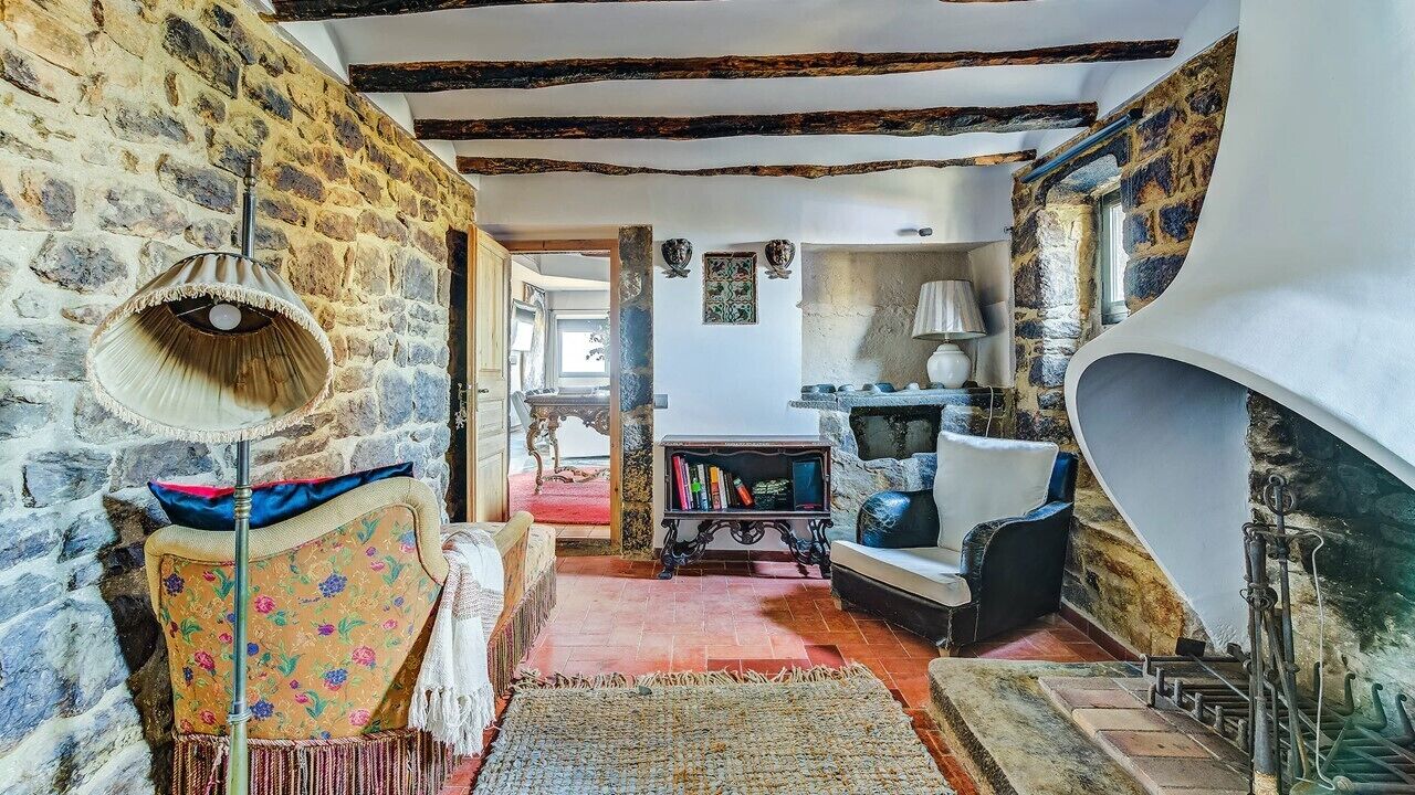 A rustic interior with exposed stone walls, a wooden ceiling, a fireplace, and a cozy seating area with a patterned armchair and a small table.