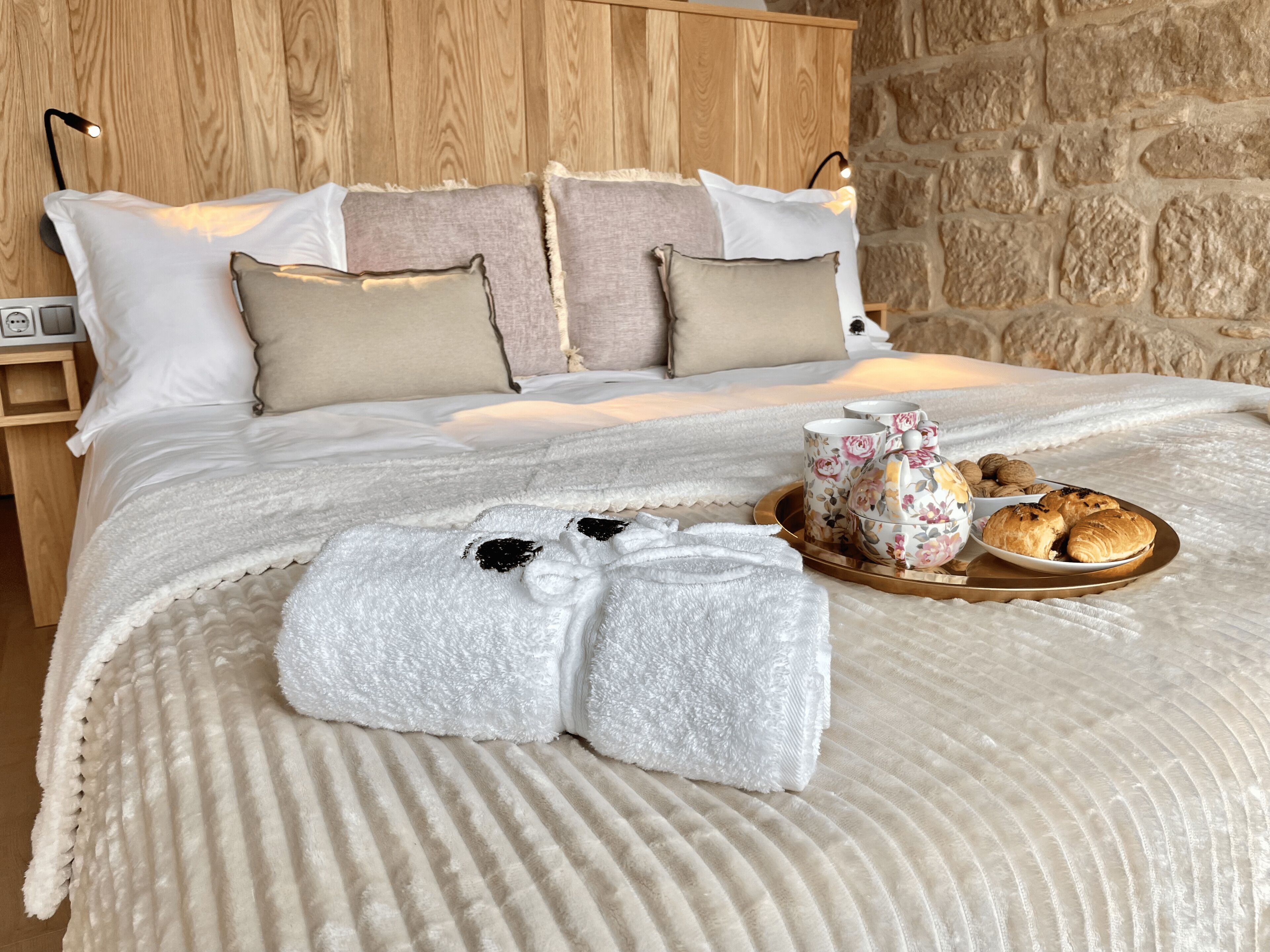 A neatly made bed with a fluffy white blanket, a set of towels, and a tray with pastries and a floral teapot.