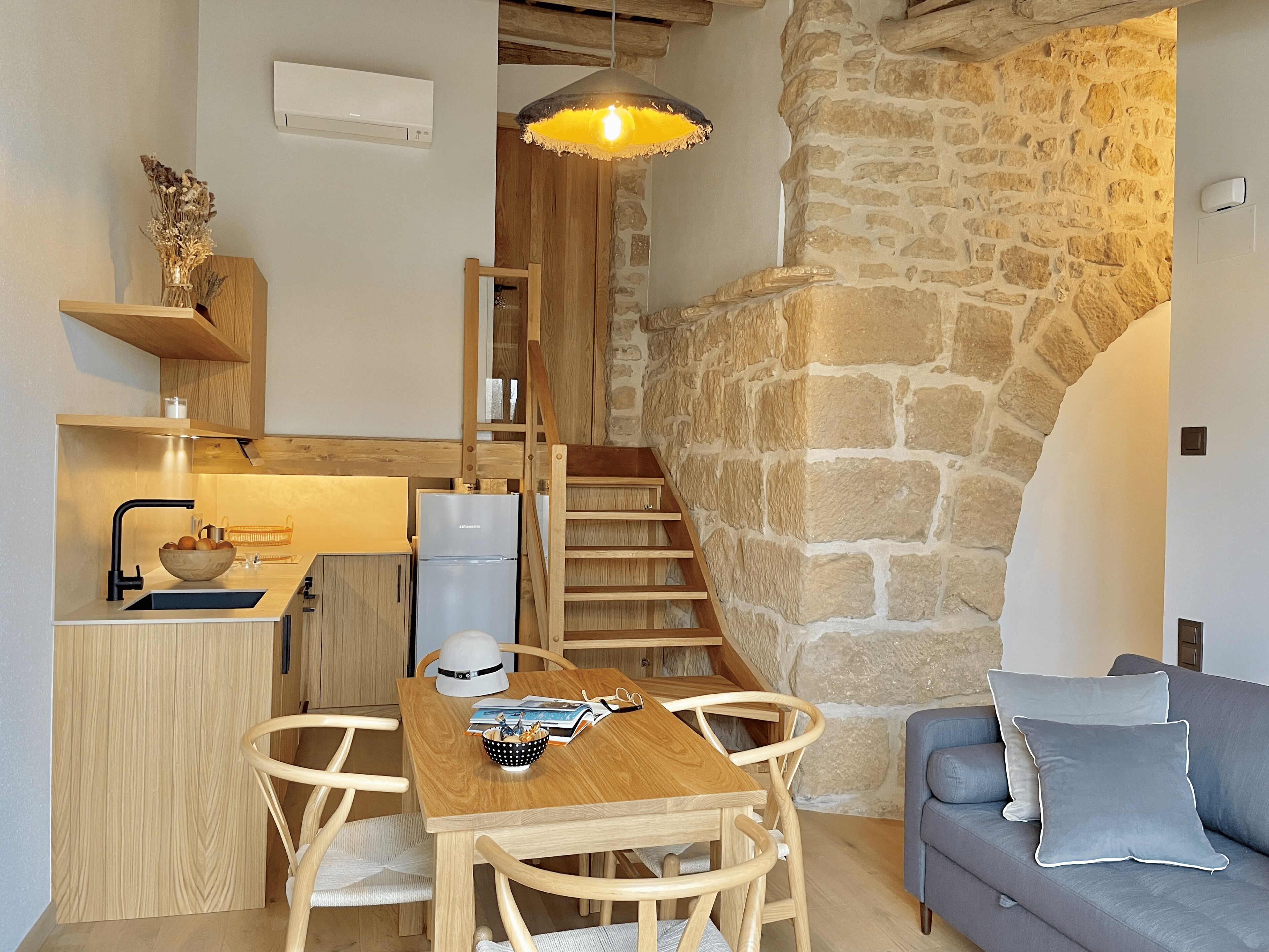 A cozy kitchen with a wooden table, chairs, and a refrigerator. A staircase with a stone wall leads to a living area with a sofa and cushions.