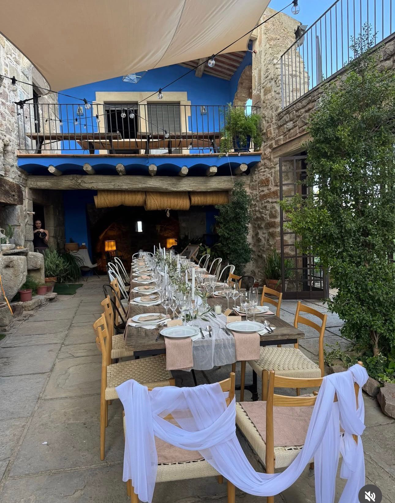 Un área de comedor al aire libre con una mesa larga dispuesta para una comida, rodeada de muros de piedra y un nivel superior azul.