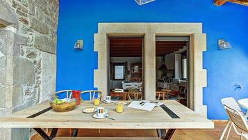 Un coin repas avec une table dressée pour deux personnes, un panier de fruits et une vue sur la cuisine par une porte ouverte.
