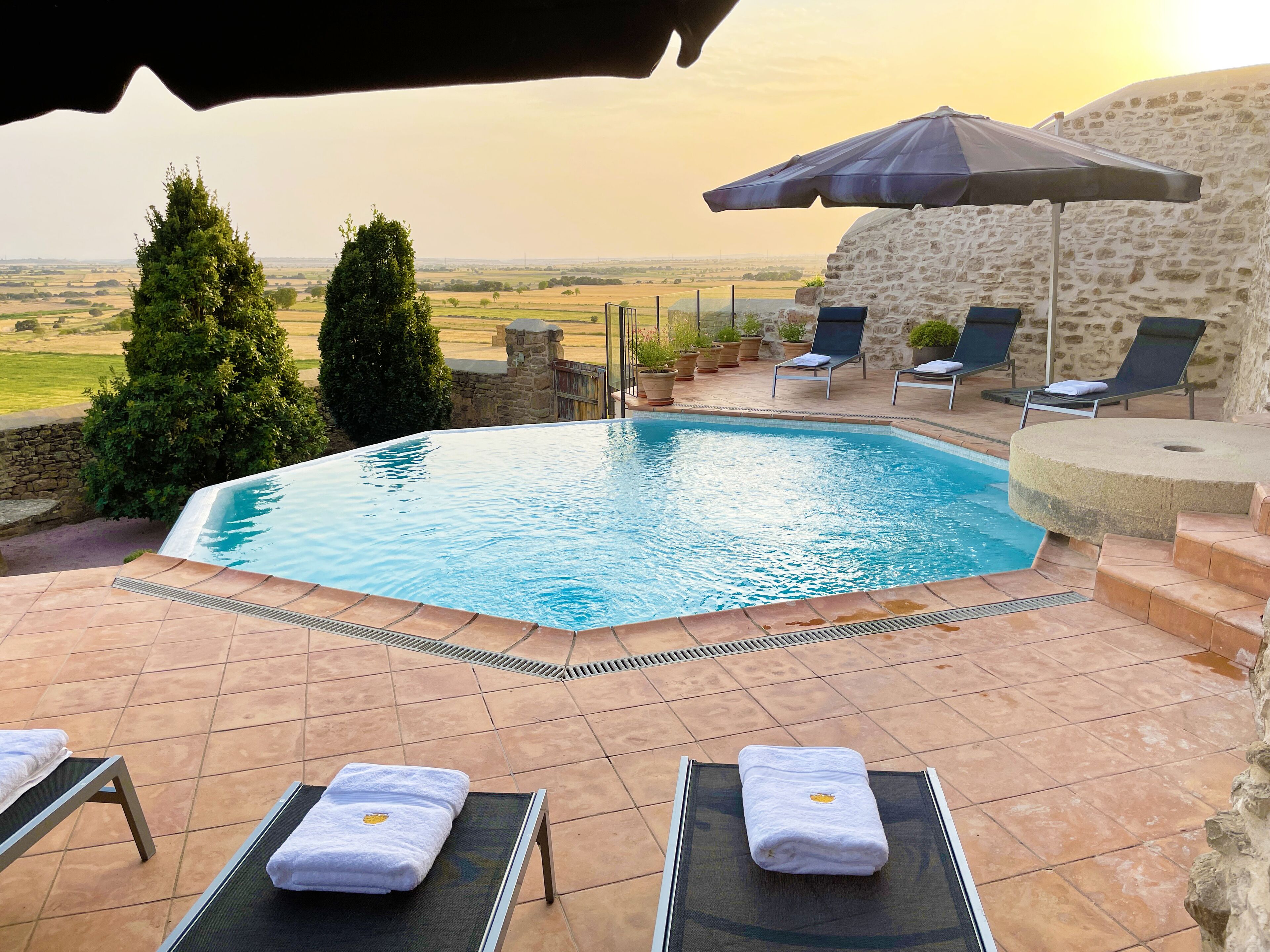 A swimming pool with lounge chairs and umbrellas, surrounded by stone walls and potted plants.