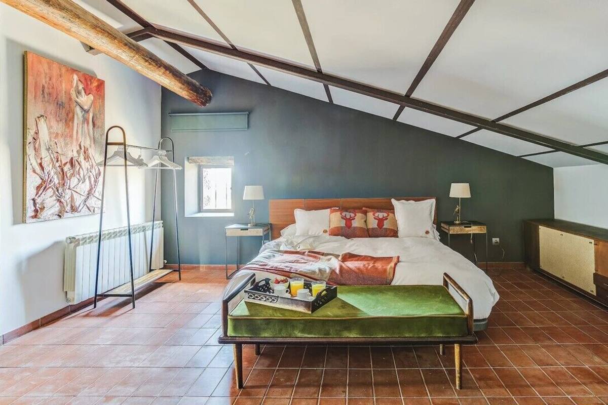 Un dormitorio con techo inclinado, una cama con almohadas y una colcha, una mesita de noche con lámpara, un radiador y un cuadro grande en la pared.