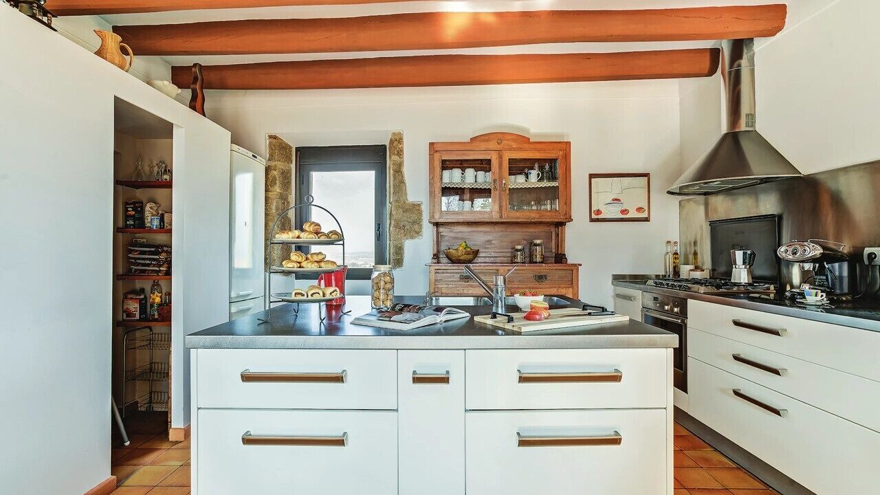 Una cocina con armarios blancos, una campana extractora de acero inoxidable y un mueble de madera con vajilla.