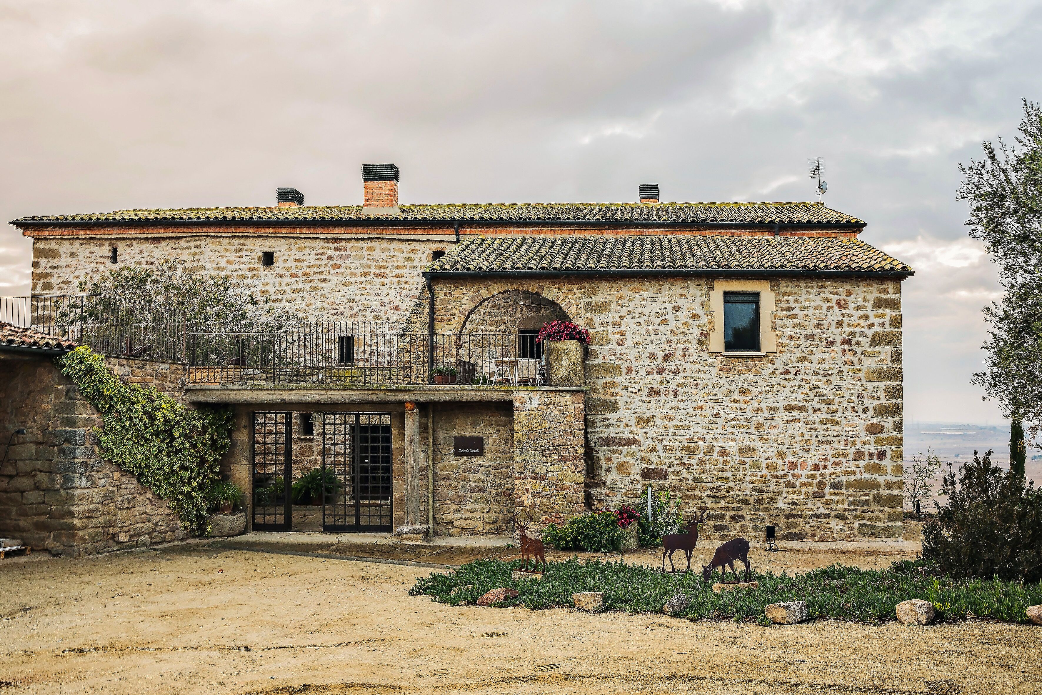 Un edificio de piedra con tejado de tejas, un balcón y un jardín con estatuas.