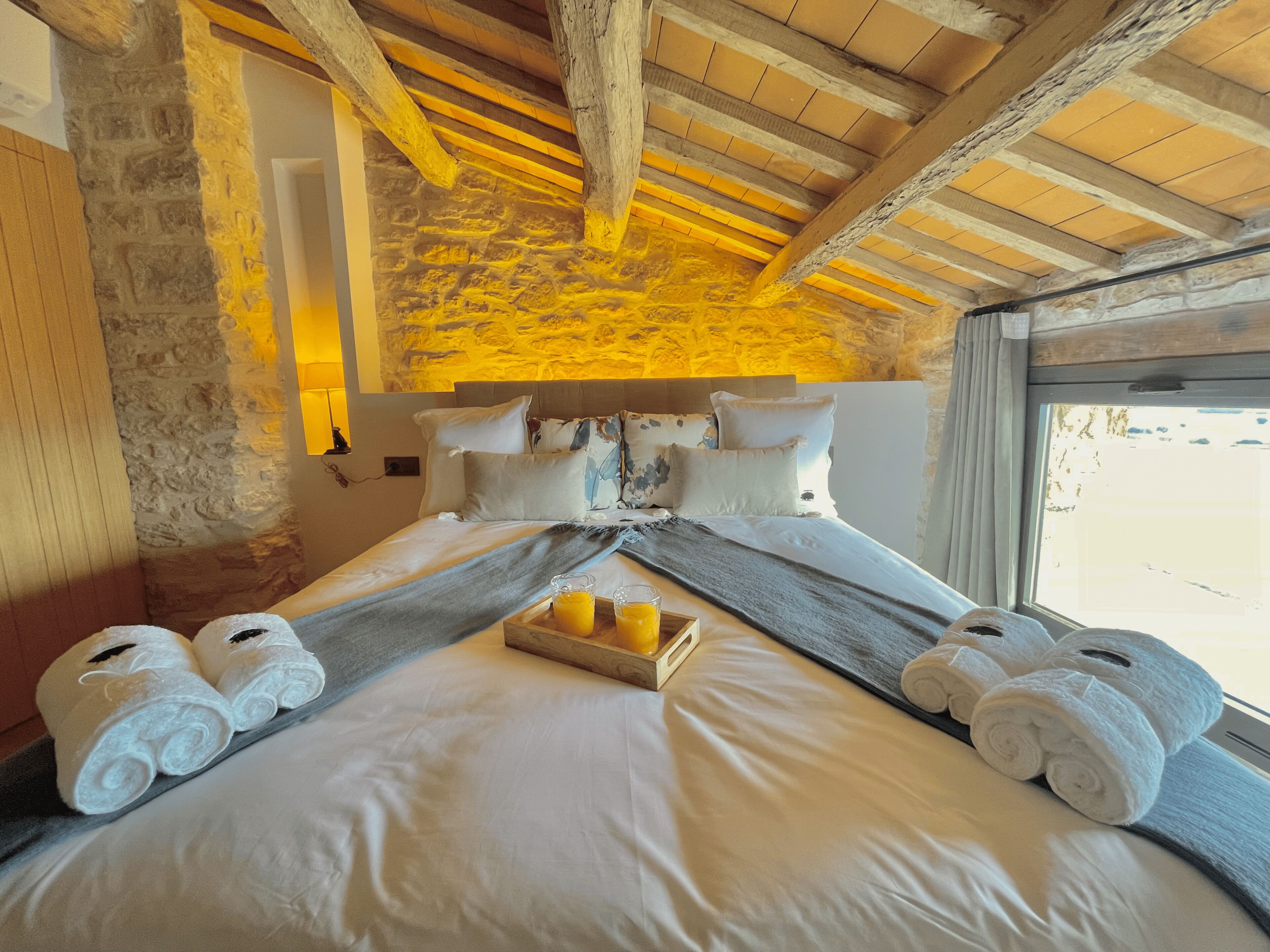 Habitación de hotel con pared de piedra, vigas de madera, cama con ropa blanca, almohadas y una bandeja con jugo de naranja.