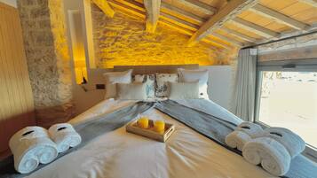 Habitación de hotel con pared de piedra, vigas de madera, cama con ropa blanca, almohadas y una bandeja con jugo de naranja.