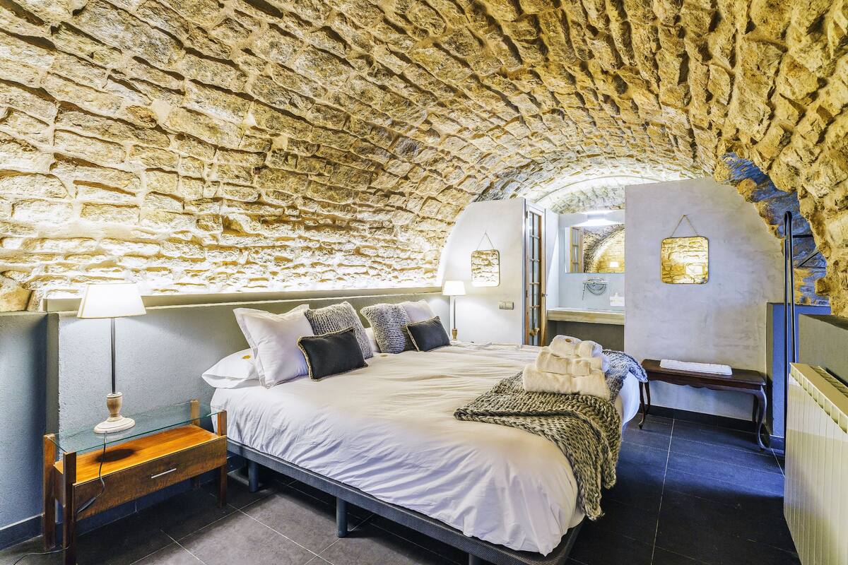 Un dormitorio con techo de piedra, cama con ropa de cama blanca, mesita de noche con lámpara y un escritorio pequeño con silla.