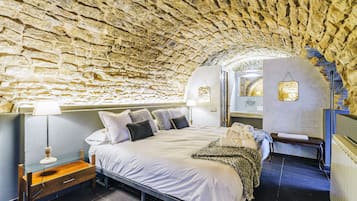 A bedroom with a stone ceiling, a bed with white bedding, a bedside table with a lamp, and a small desk with a chair.