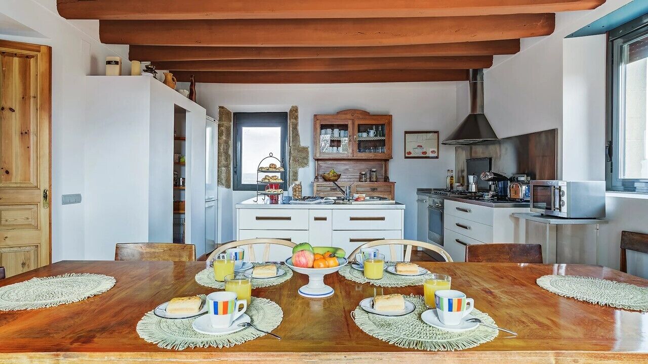 Un comedor con una mesa de madera preparada para el desayuno, una cocina al fondo y una ventana que aporta luz natural.