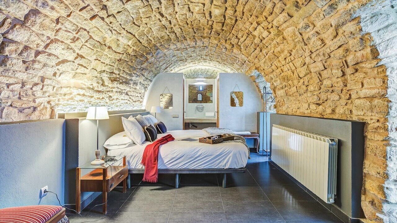 A bedroom with a stone arch ceiling, a bed with white bedding, a nightstand with a lamp, and a radiator.