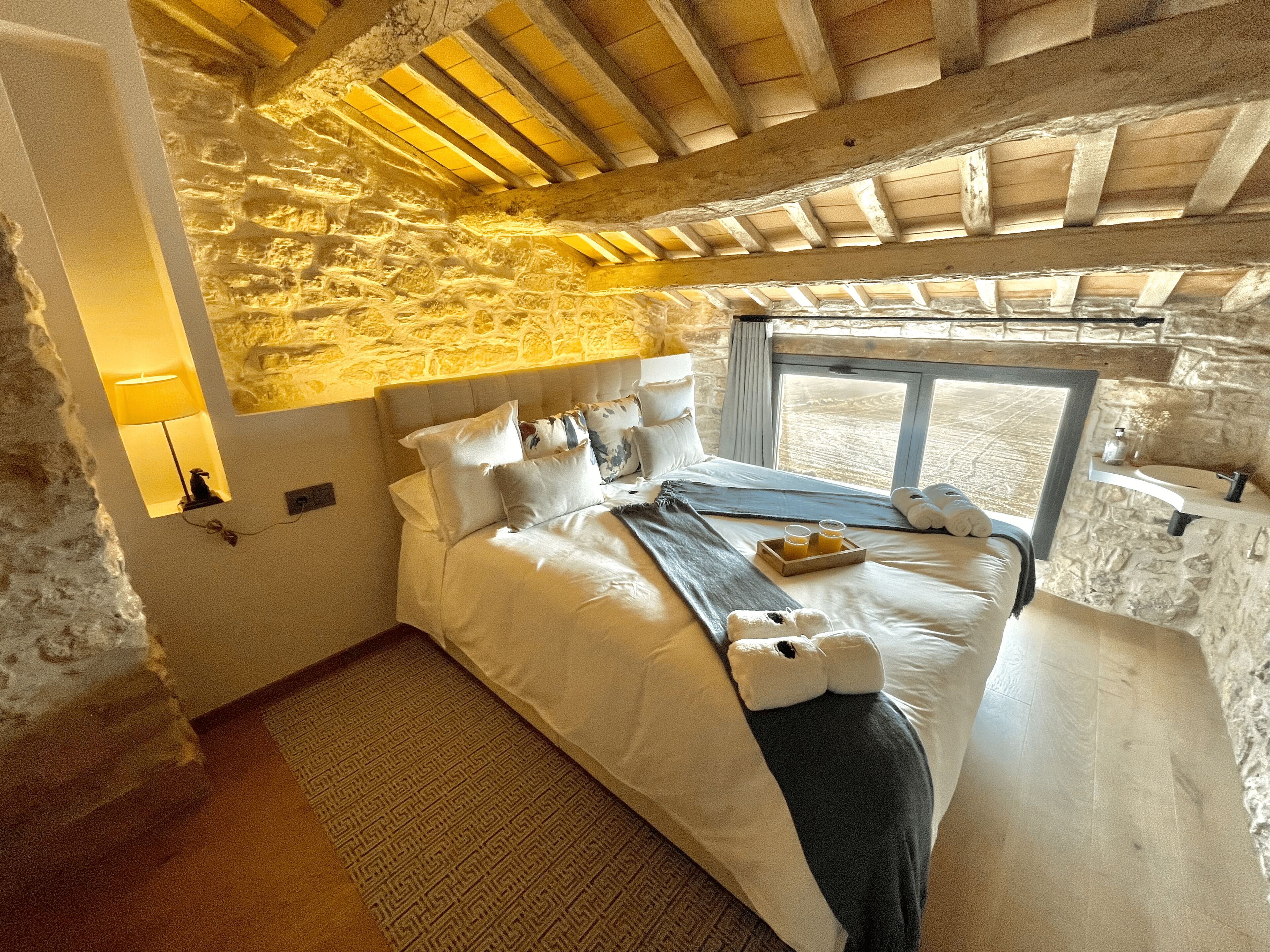 A bedroom with a stone wall, a bed with white linens, a bedside table with a lamp, and a window with a view.
