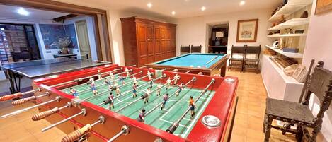 A room with a foosball table, a pool table, wooden cabinets, and a billiards table.