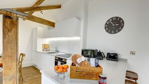 A modern kitchen with white cabinets, a wooden beam ceiling, a wall clock, a toaster, a kettle, and a bread box with oranges.