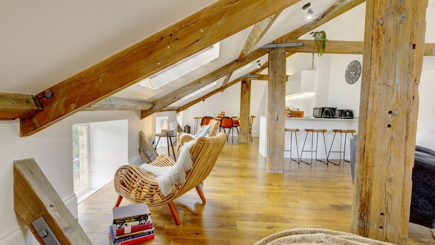 A spacious loft with wooden beams, a dining area with wicker chairs, a kitchen with bar stools, and a cozy seating area with a patterned rug.