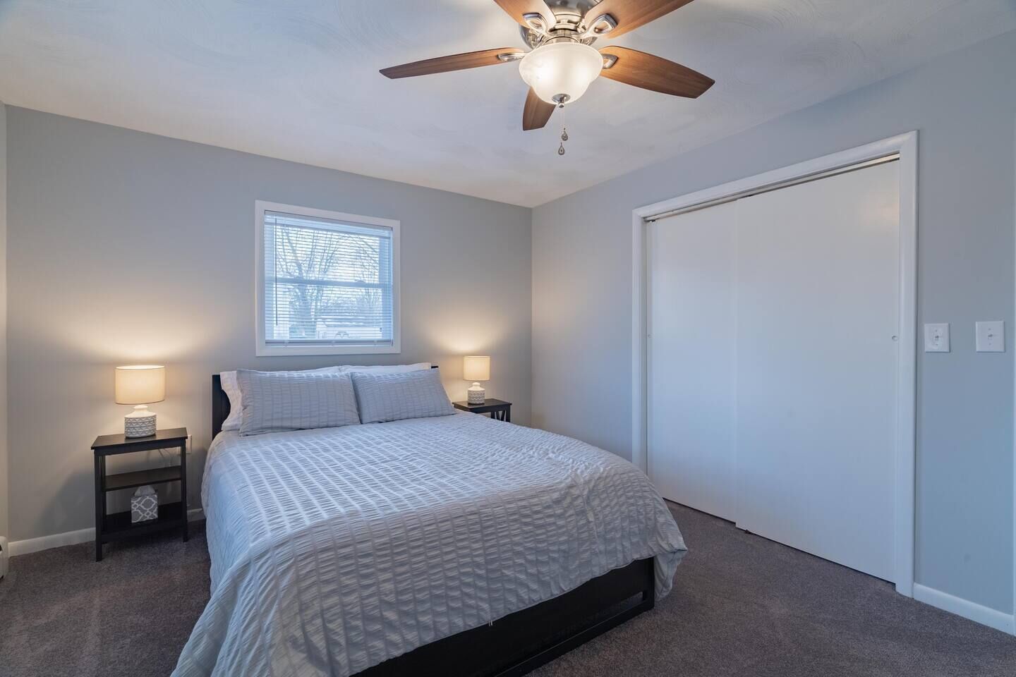 A bedroom with a bed, two bedside tables with lamps, a ceiling fan, a window with blinds, and a closed door.