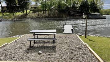 Een picknicktafel en bankje op een grindpad, naast een rustig wateroppervlak, met een steiger die in het water uitloopt en een boot in de buurt.