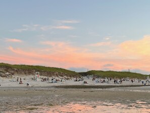 A beach with people relaxing and sunbathing, a lifeguard tower, and dunes in the background.