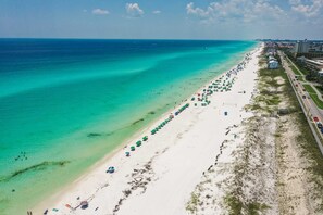 Eine Luftaufnahme eines Strandes mit einer weiten, hellen Sandfläche, klarem türkisblauem Wasser und einer Reihe von Strandsonnenliegen am Ufer.