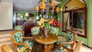 Un coin repas avec une table ronde en bois, entourée de chaises en osier ornées de motifs de feuilles vertes, un lustre avec des lampes orange et un grand vase rempli de fleurs.